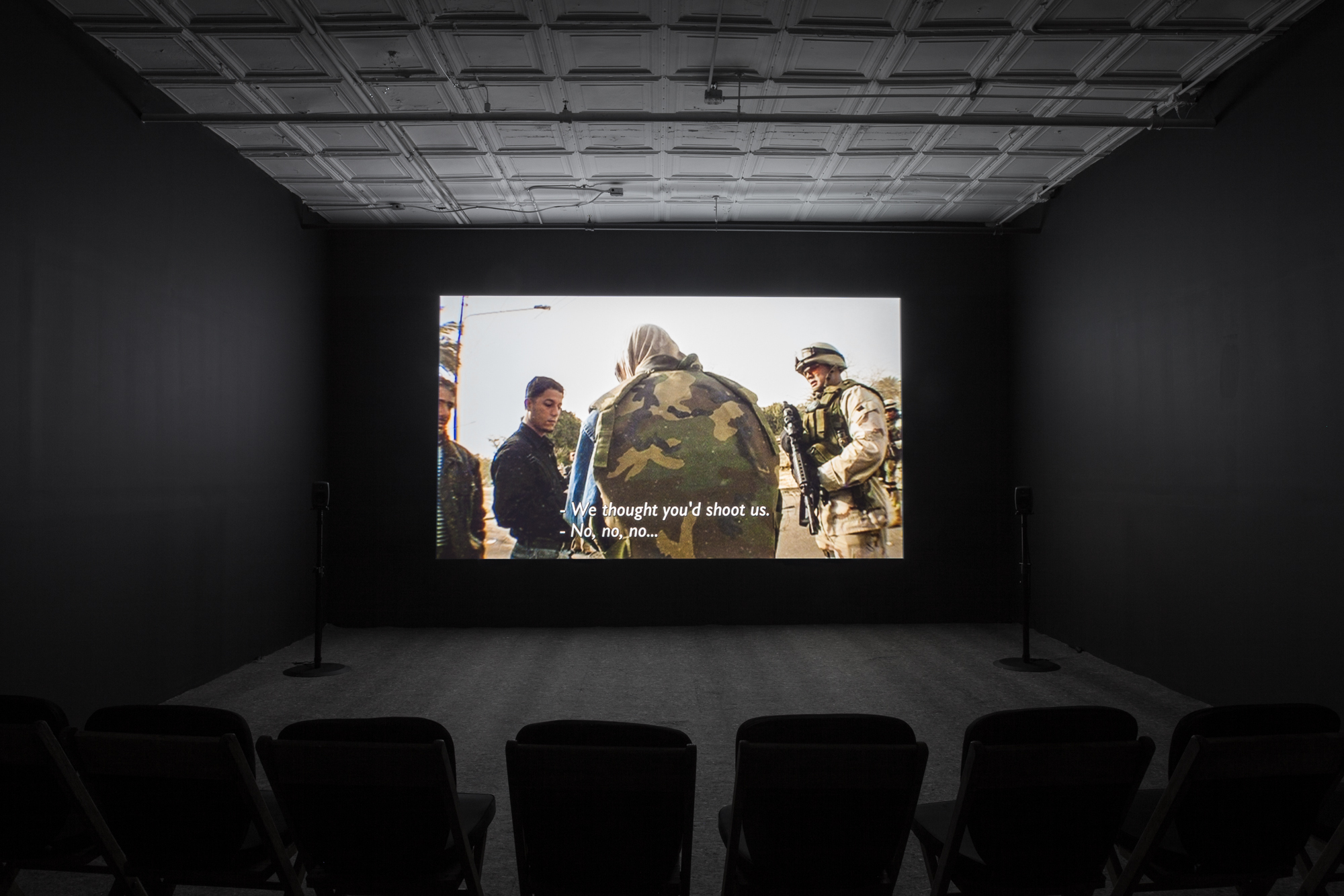 A photograph of an empty room, dimly lit with chairs in the background, where a projection is playing on the wall. The projection displays an image a soldier interacting with civilians. The caption reads, 