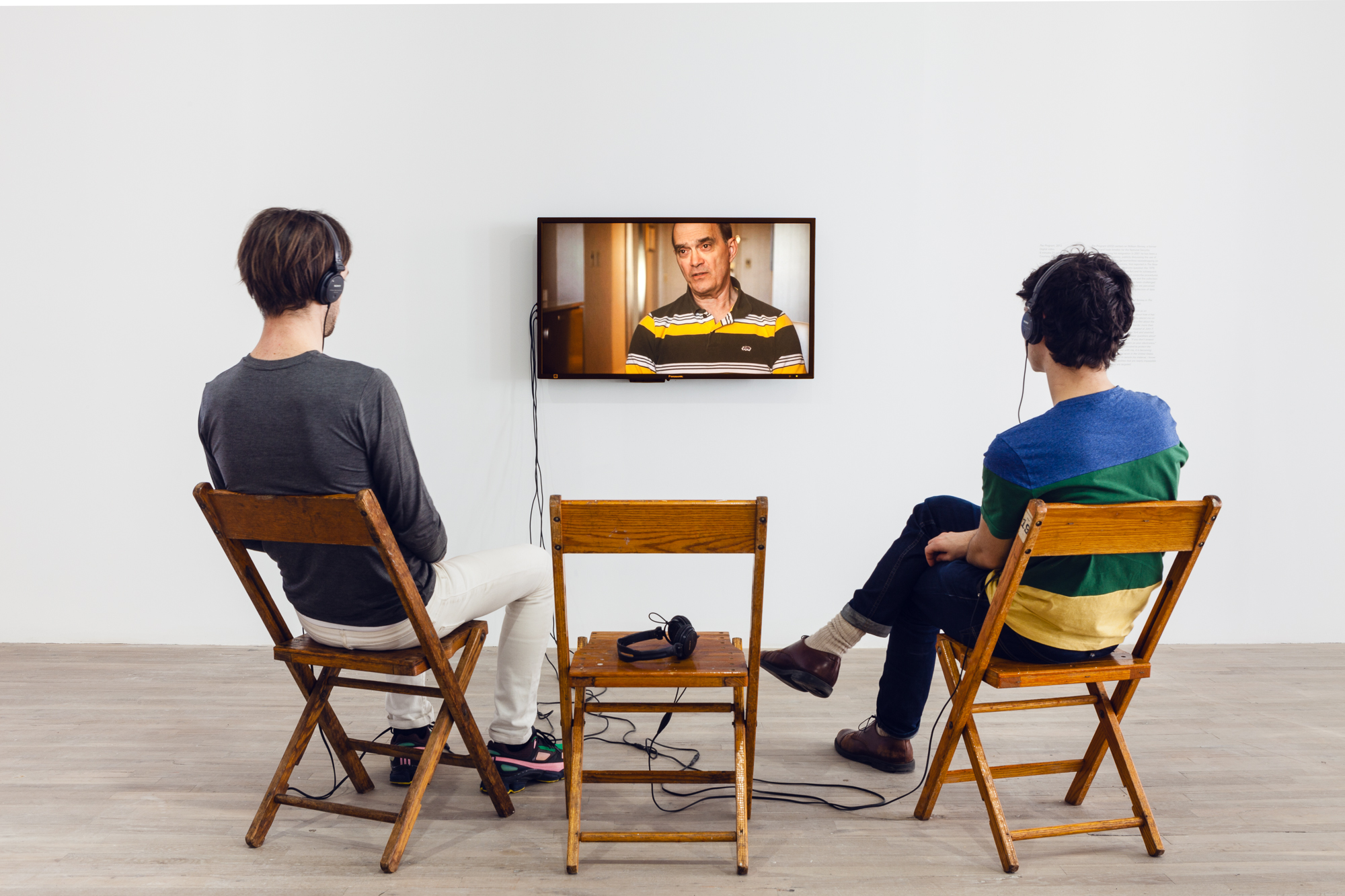 Two people wearing headphones sit in three wooden chairs arranged in front of a TV monitor, where a person wearing a black and yellow polo speaks to the camera.