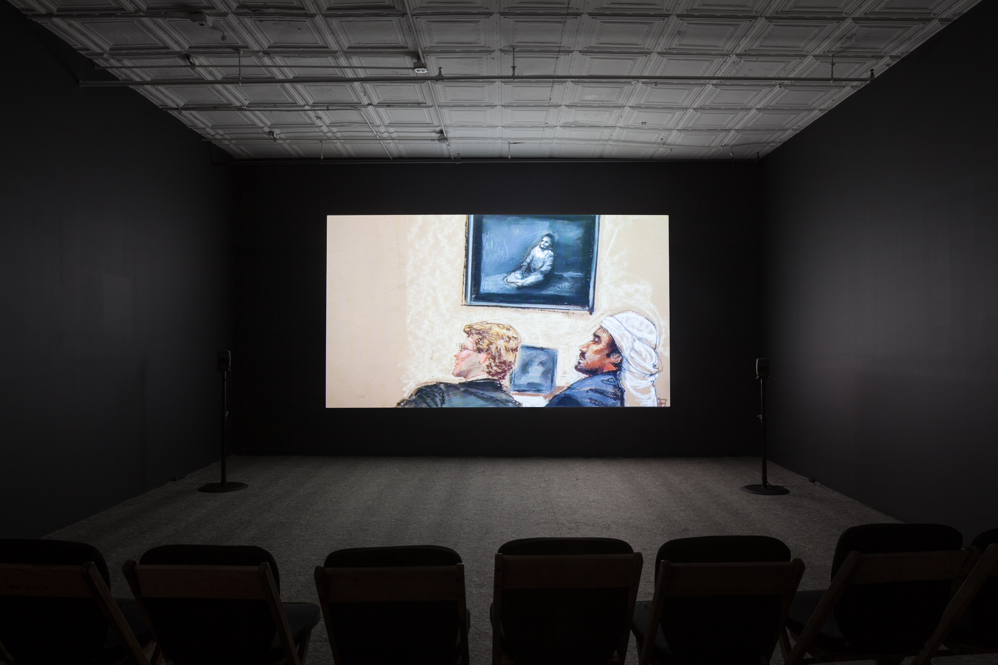 A photograph of an empty room, dimly lit with chairs in the background, where a projection is playing on the wall. The projection displays a courtroom depiction of two individuals, one with blonde hair and fair skin wearing a suit, the other tan with dark facial hair wearing a keffiyeh. Behind the seated men is an image of a man sitting on the floor, in what appears to be solitary confinement.