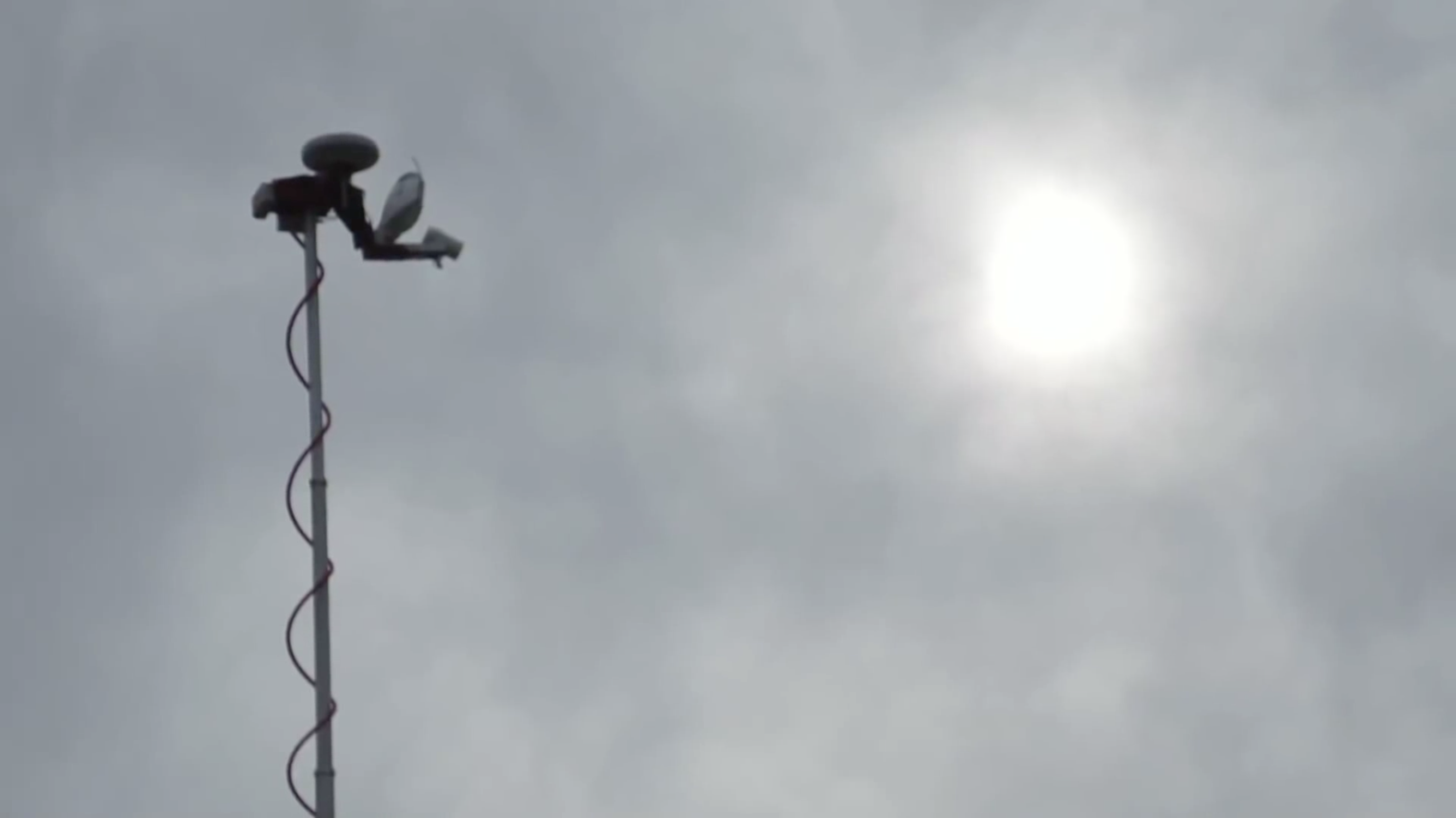 A lamp pole, left of center, in an upward angled view of a sunny, cloud covered sky. The pole has a coiled wire running upward to a security camera mounted on its top.