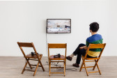 A person sits on a wooden chair where there are three arranged, wearing headphones and looks at a TV monitor. The monitor displays a construction landscape in the Middle East.