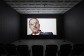 A photograph of an empty room, dimly lit with chairs in the background, where a projection is playing on the wall. The projection displays an image of a man with tan skin and facial hair, wearing a aviator-style glasses. The caption underneath his image reads, 