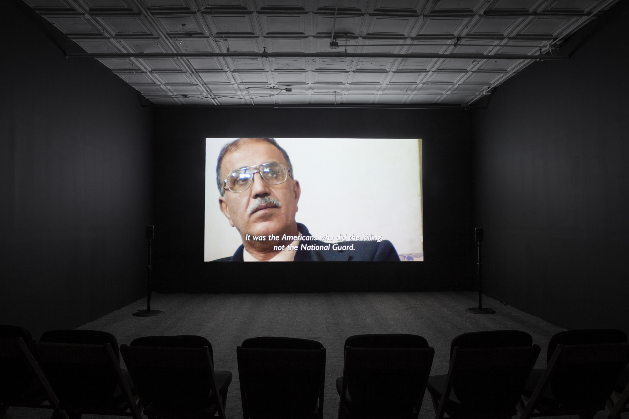 A photograph of an empty room, dimly lit with chairs in the background, where a projection is playing on the wall. The projection displays an image of a man with tan skin and facial hair, wearing a aviator-style glasses. The caption underneath his image reads, 