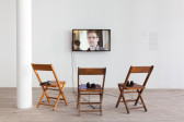 An individual TV monitor with three wooden chairs placed in front of it, each holding headphones. Edward Snowden appears speaking to the camera onscreen.