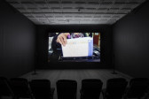 A dimly lit, empty room filled with several low chairs. A projection illuminates a dark wall opposite the chairs. The projection displays an image of a figure, their index finger blotted purple, inserting a document written in Arabic into a machine.