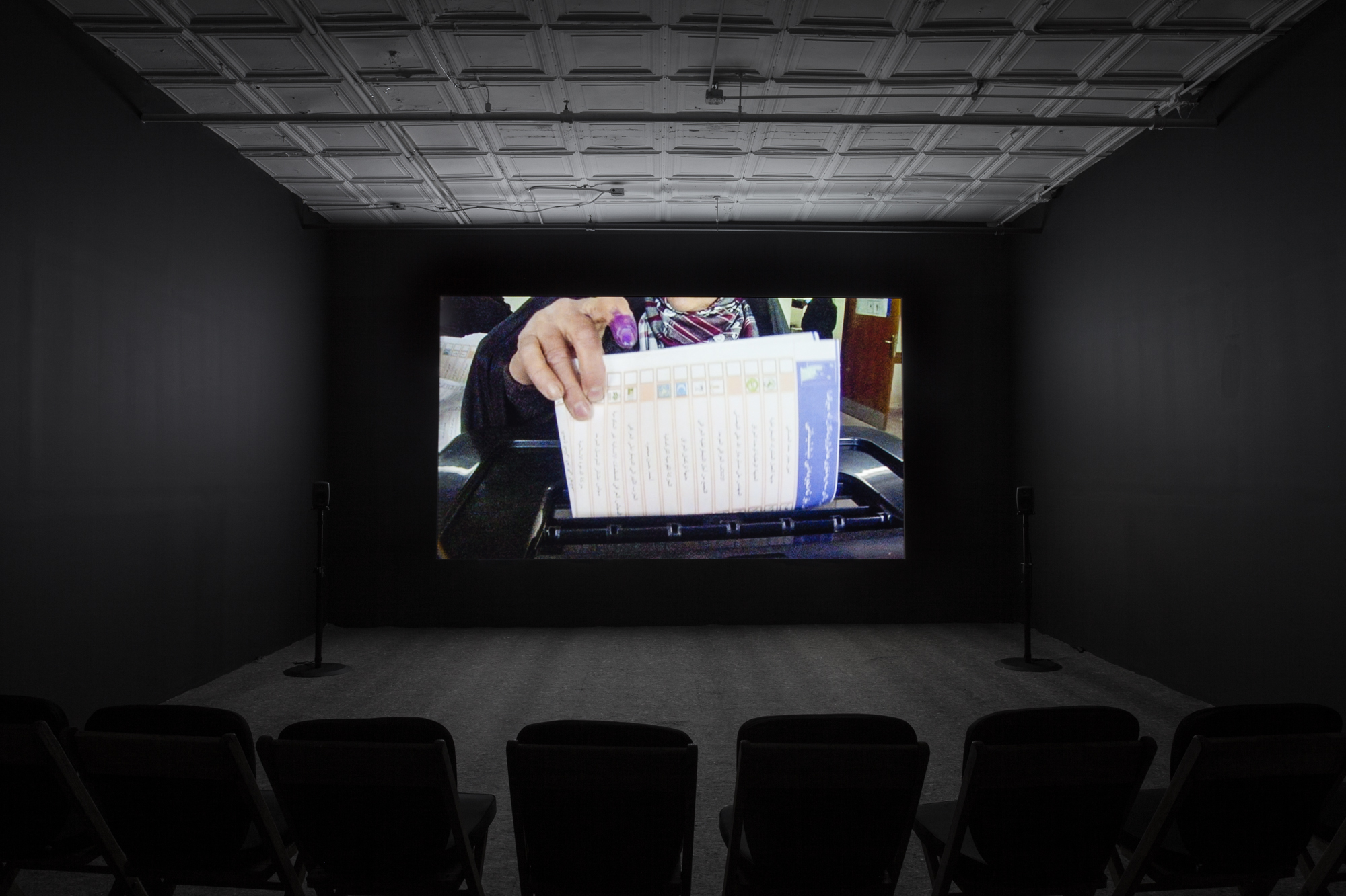 A dimly lit, empty room filled with several low chairs. A projection illuminates a dark wall opposite the chairs. The projection displays an image of a figure, their index finger blotted purple, inserting a document written in Arabic into a machine.