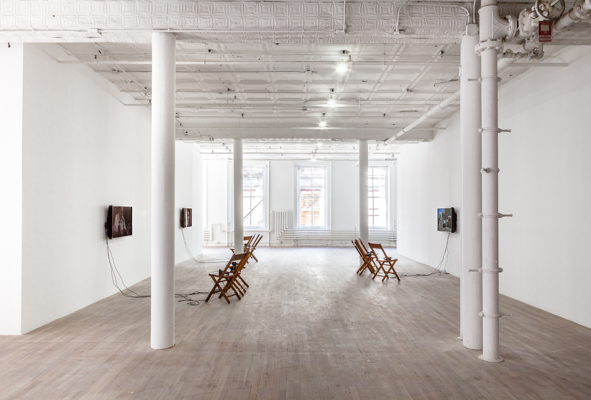 Several wooden chairs placed in front of three monitors installed on two walls of an exhibition space. Each of the monitors displays a film.