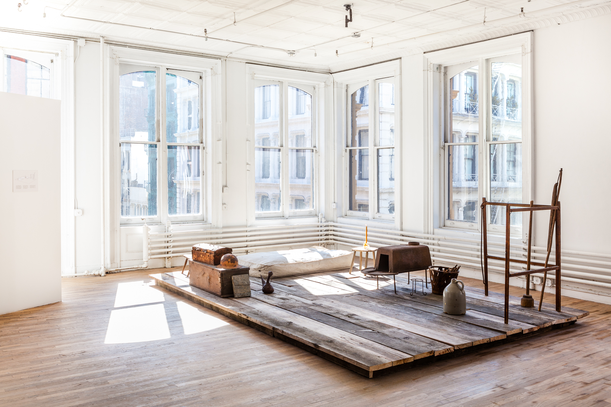 A section of wooden flooring is displayed on the ground near four windows on adjacent walls of an exhibition space. A small bed, a candle on a night table, a metal stove, and a several chests stacked on top of one another are arranged on the wooden boards.