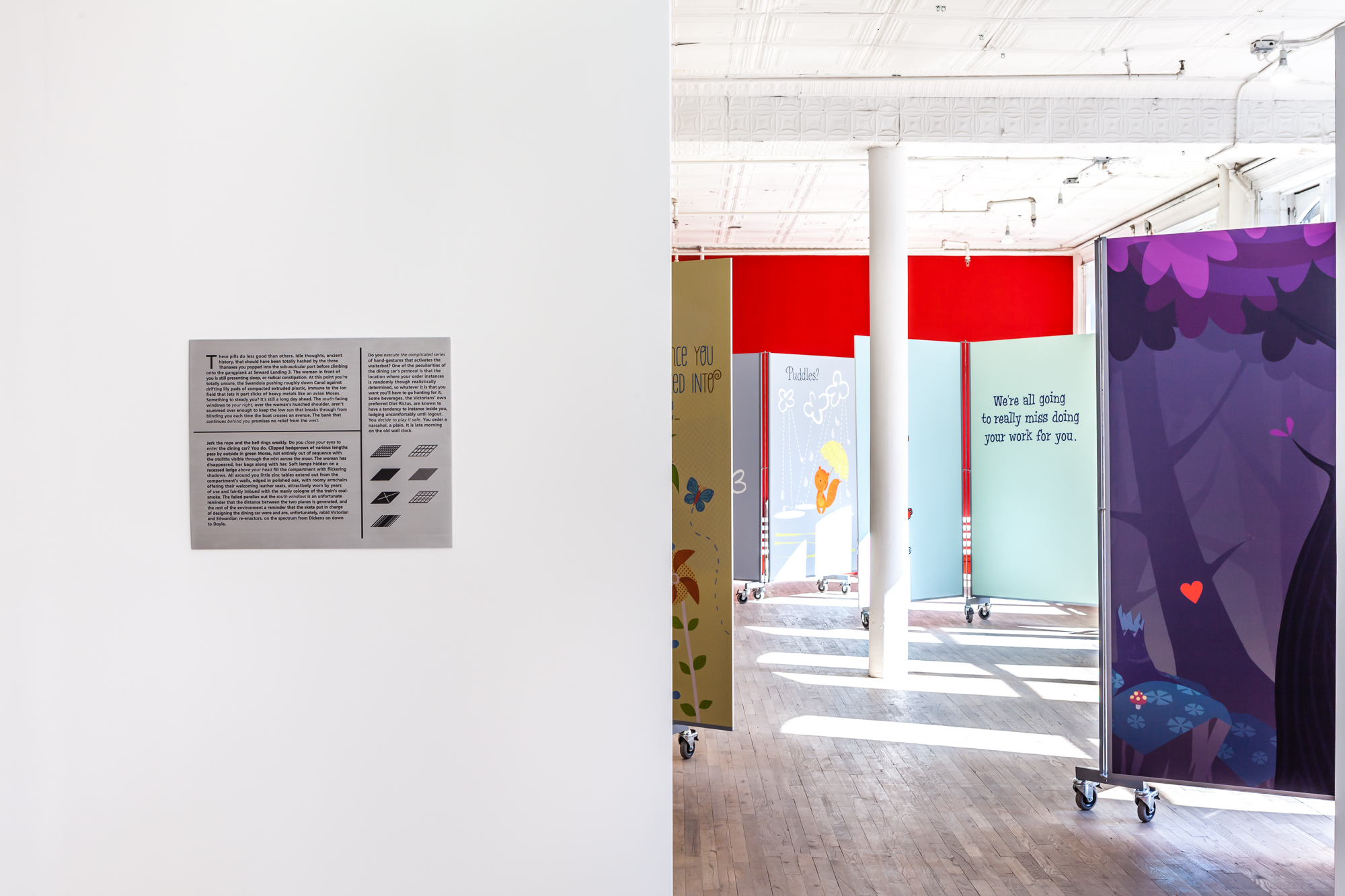 A metal plaque engraved with text is displayed on a white gallery wall that extends into the left half of the image's frame. Past and to the right of the wall, a series of colorful panels are staggered around a white column in front of a red wall. Some of the panels are decorated with cartoonish illustrations of animals and forests. Large text on one of the visible panels reads, 