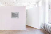 A freestanding white wall displaying a metal plaque engraved with paragraphs of text juts in front of another angled, white wall in an exhibition space. A large paragraph of wall vinyl text is displayed on the second, angled wall.