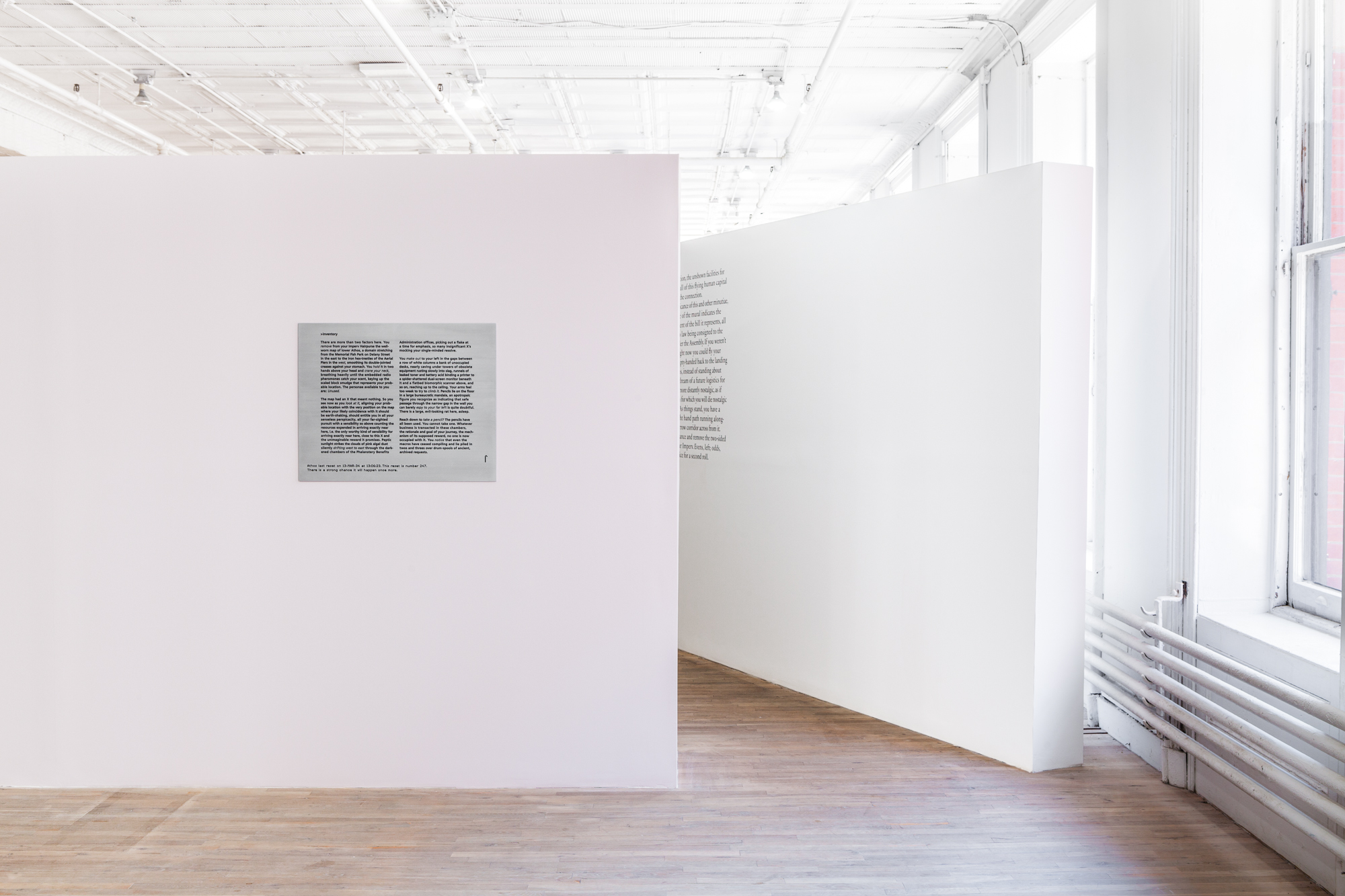 A freestanding white wall displaying a metal plaque engraved with paragraphs of text juts in front of another angled, white wall in an exhibition space. A large paragraph of wall vinyl text is displayed on the second, angled wall.