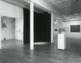 Black and white image of several sculptures and two-dimensional artworks installed in a gallery space bisected by three white columns. Behind the columns, a large dark tarp covers two gallery walls that form a small corner. To the right of the columns, a large glass container filled with honey rests on a plinth. To the left of the columns, a wide doorway opens up into another gallery room, where several chandelier-like sculptures hang from the ceiling, while three spindly structures are propped on the floor.