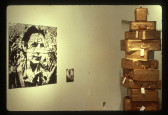 A fractured image of a person hung on a white wall next to a tall stack of suitcases painted gold.