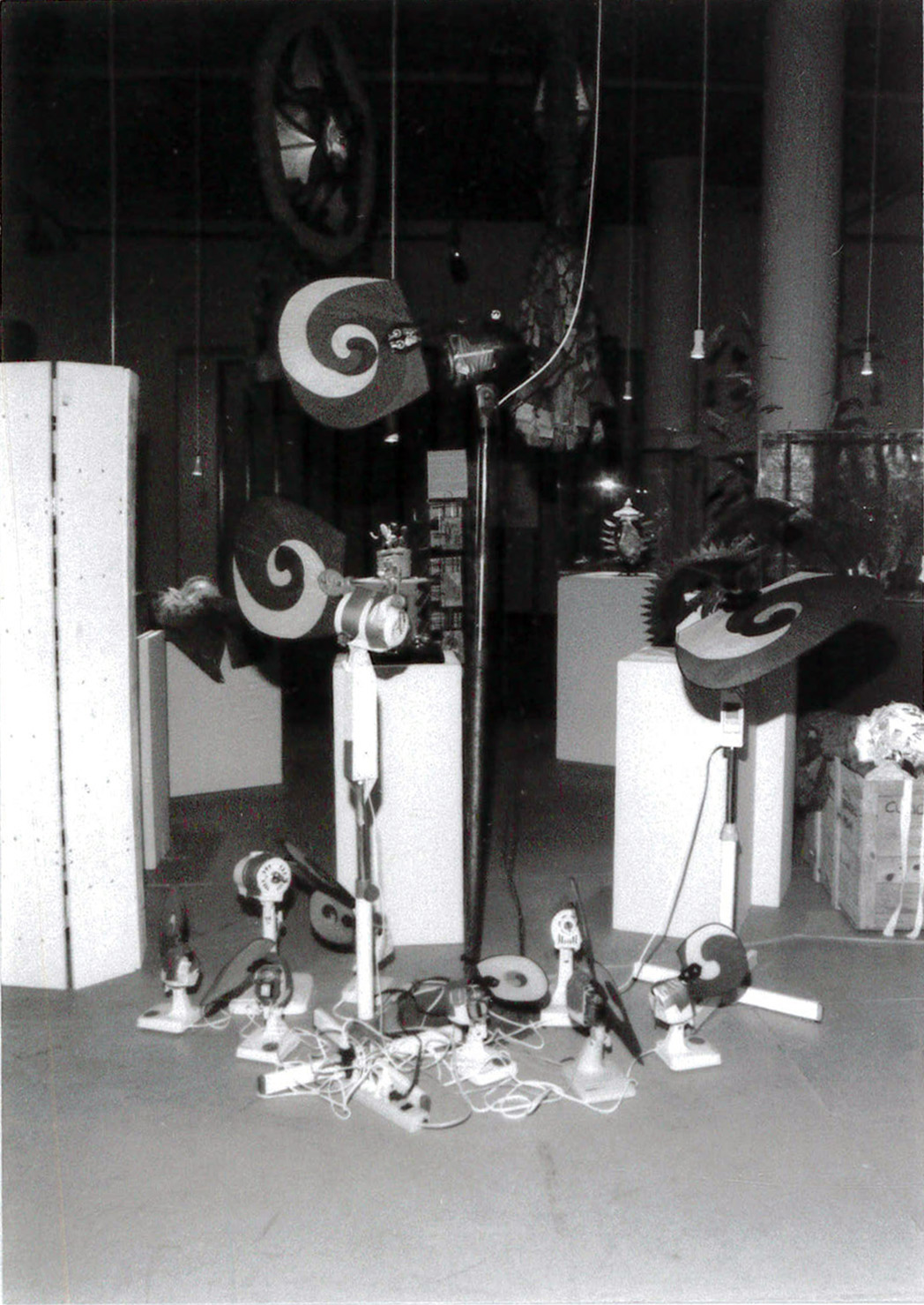 A black and white photograph of a room filled with plinths of varying heights, several objects suspended from the ceiling, and a pile of small fans with a mess of wires sitting on the floor.
