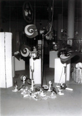 A black and white photograph of a room filled with plinths of varying heights, several objects suspended from the ceiling, and a pile of small fans with a mess of wires sitting on the floor.