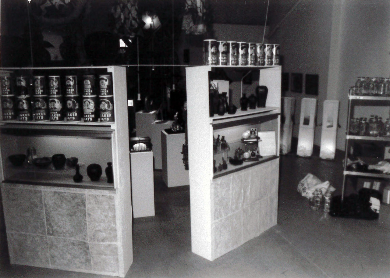 A black and white photograph of two shelves, each with several Quaker Oats canisters and vases of assorted shapes. Beyond the shelves are several plinths housing objects, and a metal rack with dozens of glass jars.