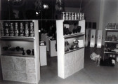 A black and white photograph of two shelves, each with several Quaker Oats canisters and vases of assorted shapes. Beyond the shelves are several plinths housing objects, and a metal rack with dozens of glass jars.