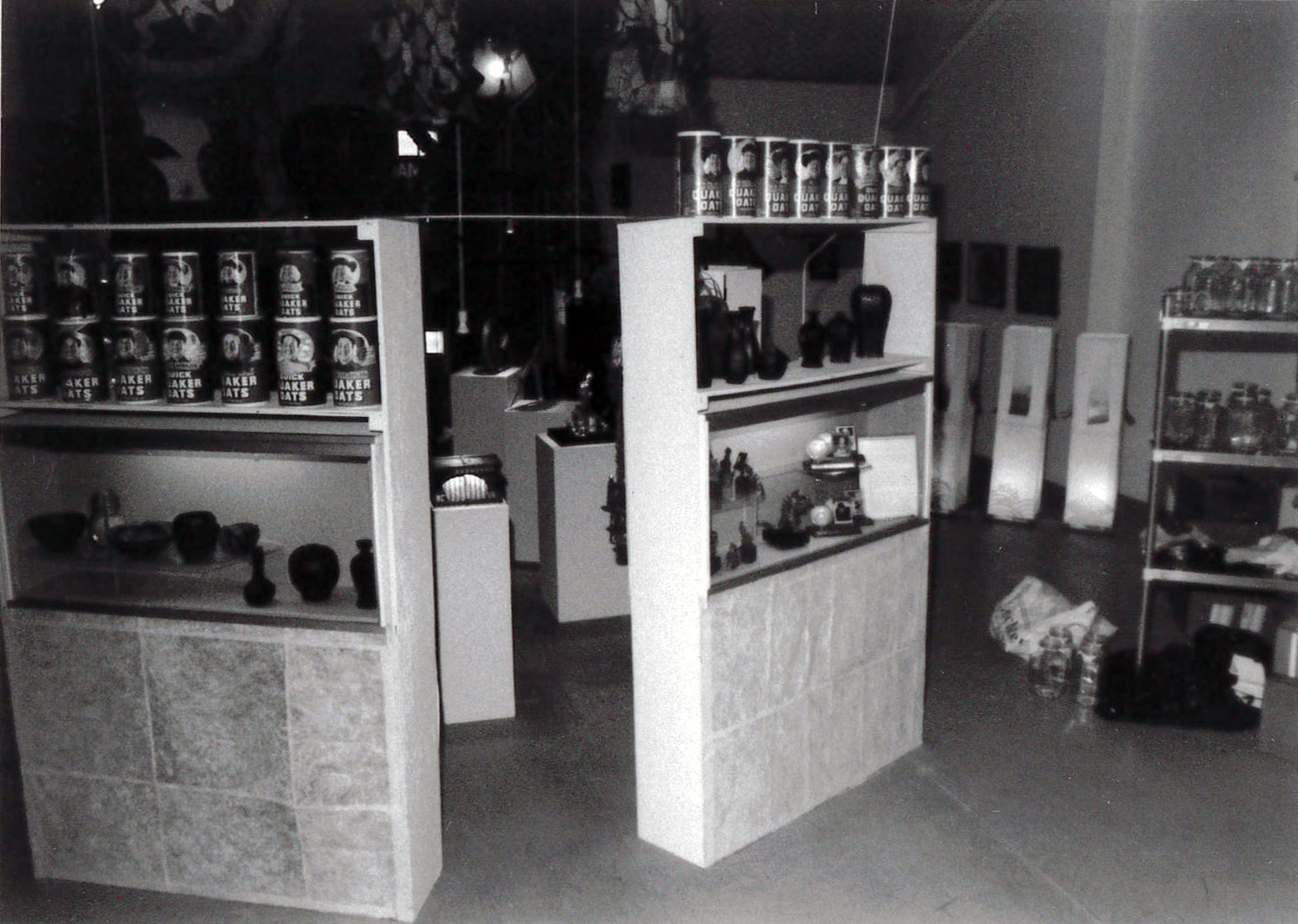 A black and white photograph of two shelves, each with several Quaker Oats canisters and vases of assorted shapes. Beyond the shelves are several plinths housing objects, and a metal rack with dozens of glass jars.