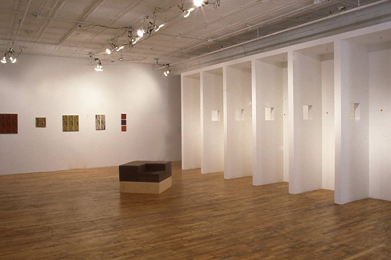 A gallery space with a seat made from a wooden block in the center. On one wall, five artworks hang in a horizontal line. Several perpendicular walls emerge from the gallery wall to the right of the space. The perpendicular walls are arranged such that they create small booths, with a square hole in each wall.