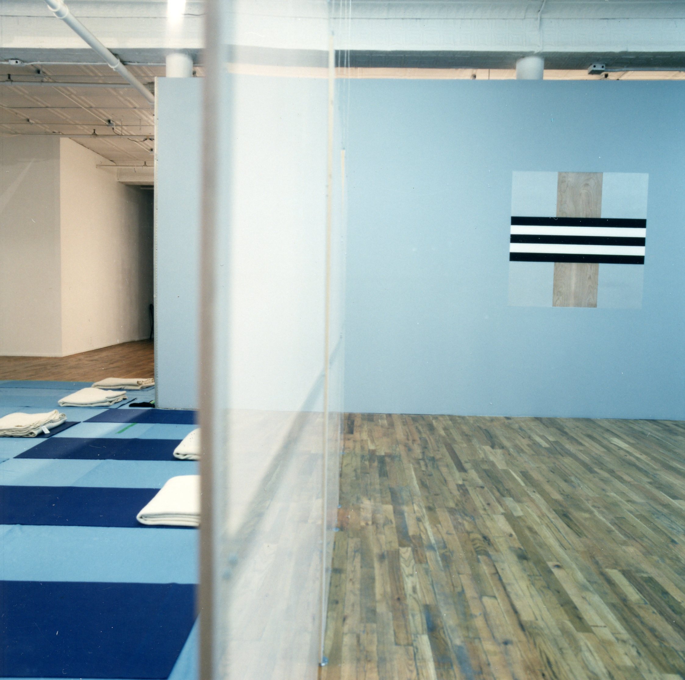 A room divided by a glass partition. On the righthand side, the walls are blue with a wooden floor, and a square artwork painted with stripes hangs on the wall. On the lefthand side, the floor is light blue and covered with dark blue mats placed at even intervals. A white, folded towel is placed on each of the mats.
