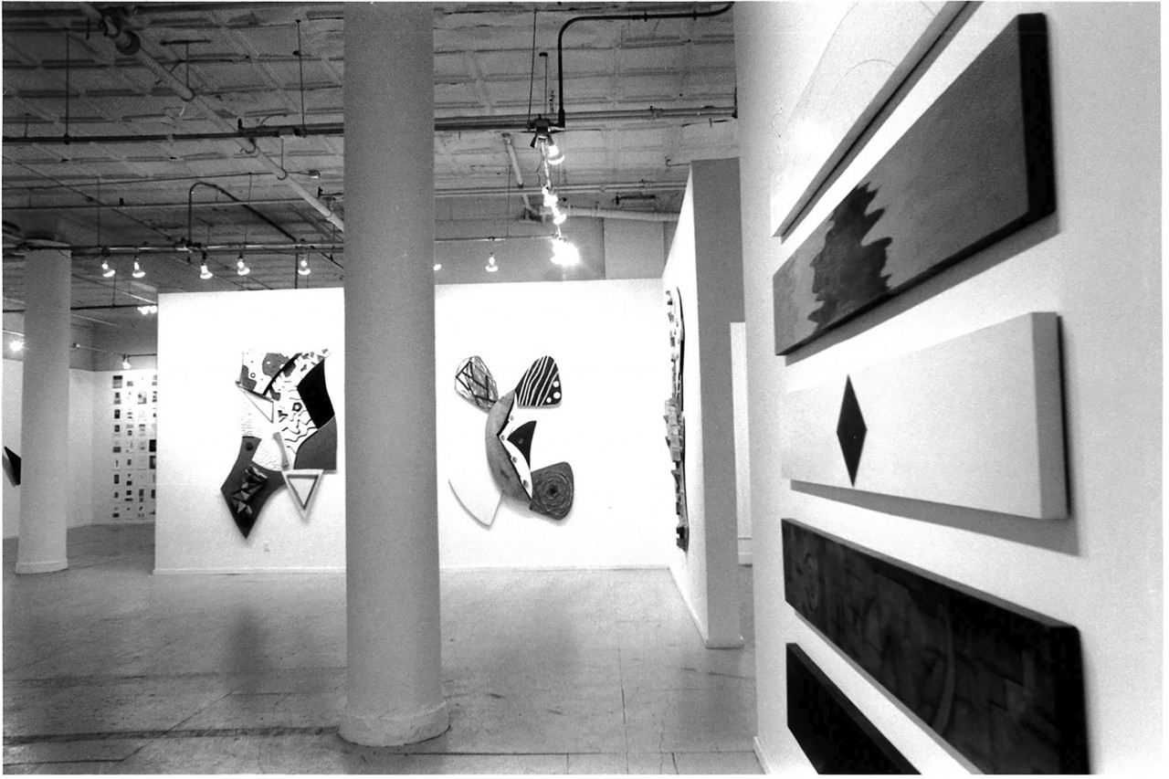 On the right abstract paintings on rectangular wooden blocks, a column bisects the frame centrally and painted wood constructions are visible on the walls in the background in a white-walled gallery setting.