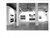 A series of small rectangular abstract paintings hang above one another on the walls of a white-walled gallery setting with two columns visible in front.