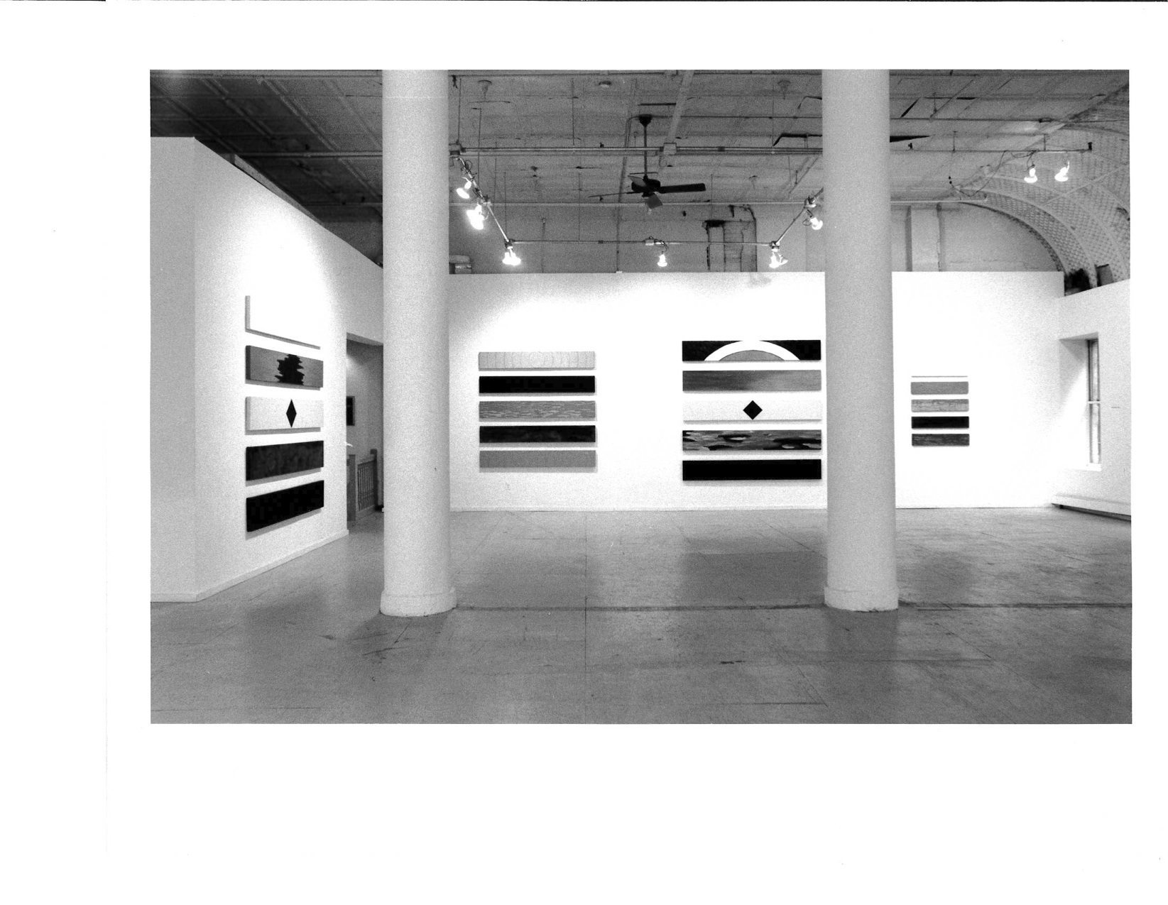 A series of small rectangular abstract paintings hang above one another on the walls of a white-walled gallery setting with two columns visible in front.