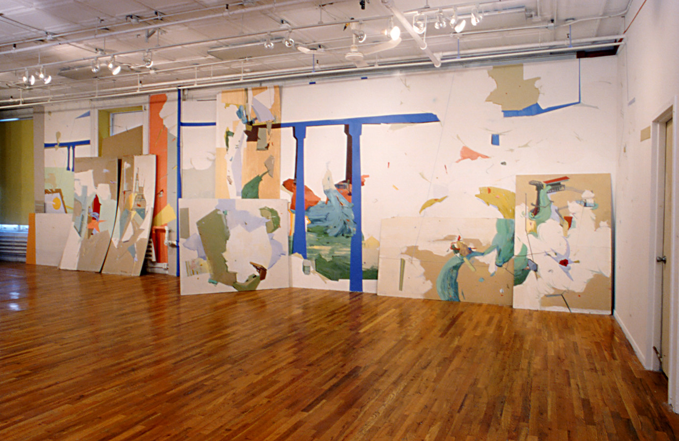 A large gallery space with white walls and wooden flooring. The length of a long gallery wall has been painted with amorphous areas of turquoise, yellow, red, and green color amidst vaguely cloud-like white shapes. Several rectangular canvases decorated with similar designs lean against the wall. A large blue shape resembling a bridge stretches across the wall design.