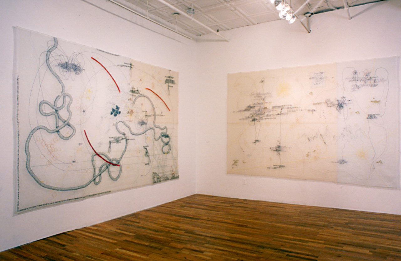 Corner of a gallery space with white walls and wooden flooring. Two large rectangular lengths of translucent, white cloth embroidered with amorphous red and blue linework are installed on the corner