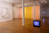 Several artworks installed in a large gallery space with white walls and wooden flooring. A television monitor rests on a low acrylic box on the floor to the room's right side. A warped purple and pink image resembling a kaleidoscopic star shape is displayed on its screen. There are white columns to the left of the monitor. Hanging on a gallery wall on the image's far left side is a large piece of translucent, white cloth embroidered with amorphous red and blue linework. The far gallery wall behind the twinned columns has been painted with slanted, striped areas of peach, yellow, and ochre color.
