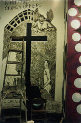 A person sits in a chair with a ladder next to them, bent over and painting on the bottom of a wall. The wall is painted top to bottom with a large black cross and several figures. Hand-painted text at the top of the wall reads, 