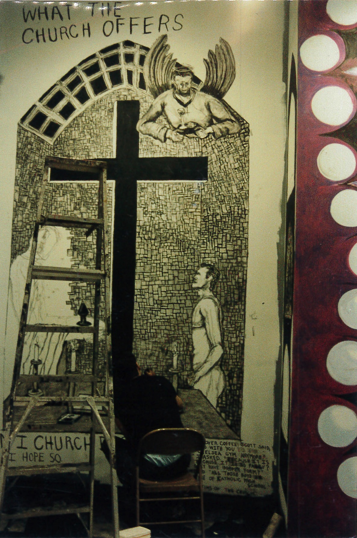 A person sits in a chair with a ladder next to them, bent over and painting on the bottom of a wall. The wall is painted top to bottom with a large black cross and several figures. Hand-painted text at the top of the wall reads, "WHAT THE CHURCH OFFERS."