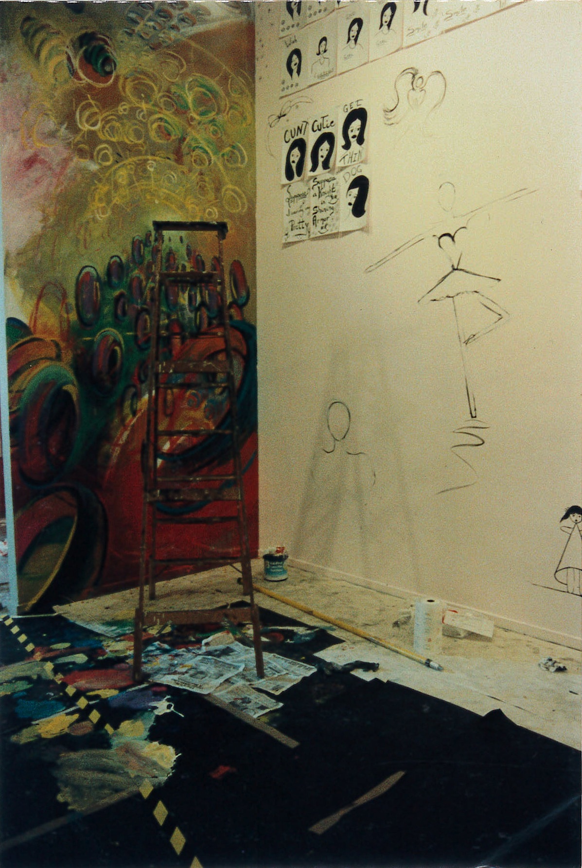 A wooden ladder standing in the corner of a room with half-finished paintings on the wall. On the left wall, there is a colorful mural composed of swirls and circles. On the other wall, there is a sketch of a ballerina with dozens of black and white portraits of a woman