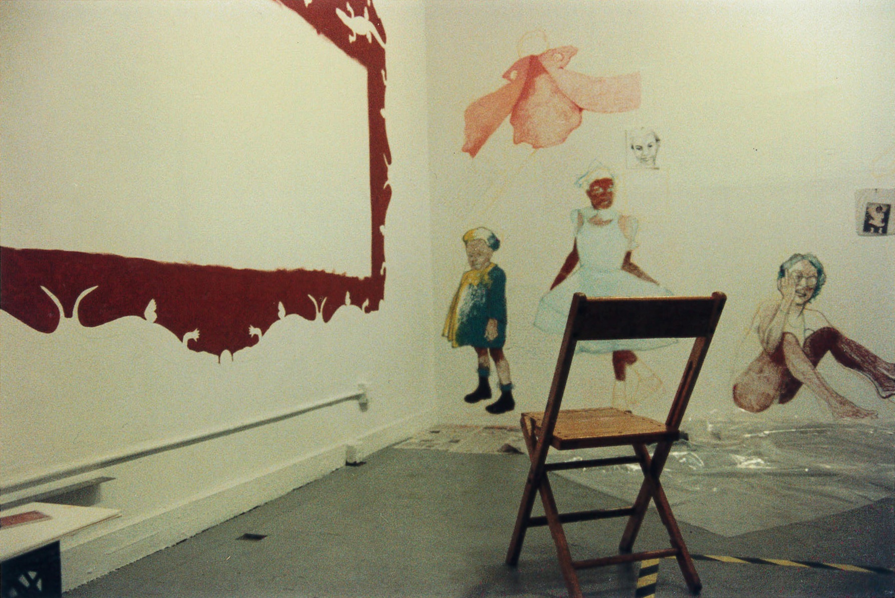 A wooden chair facing the corner of a room. Painted on one wall is an empty red frame. On the perpendicular wall, there are several half-finished paintings of children.