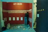 A person working in a small space enclosed by walls, painted red with white text. At the top of the wall, large letters read 
