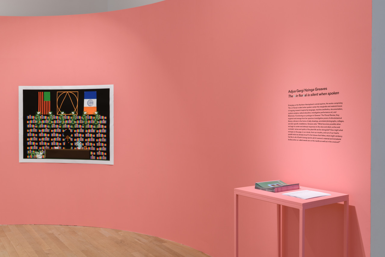 A photograph of a print mounted on a pink gallery wall next to wall text. Below the wall text is a table with papers stacked on top.