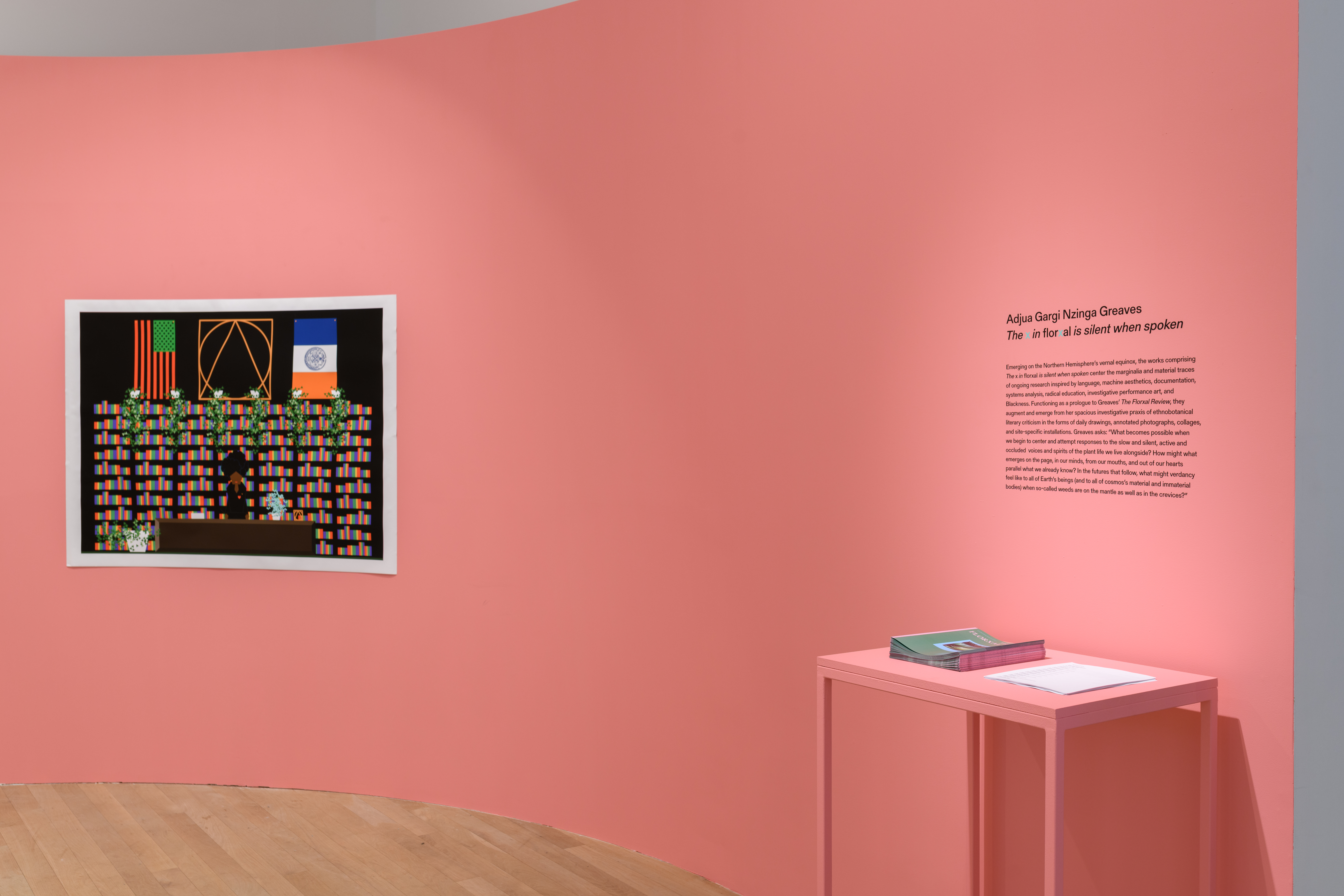 A photograph of a print mounted on a pink gallery wall next to wall text. Below the wall text is a table with papers stacked on top.