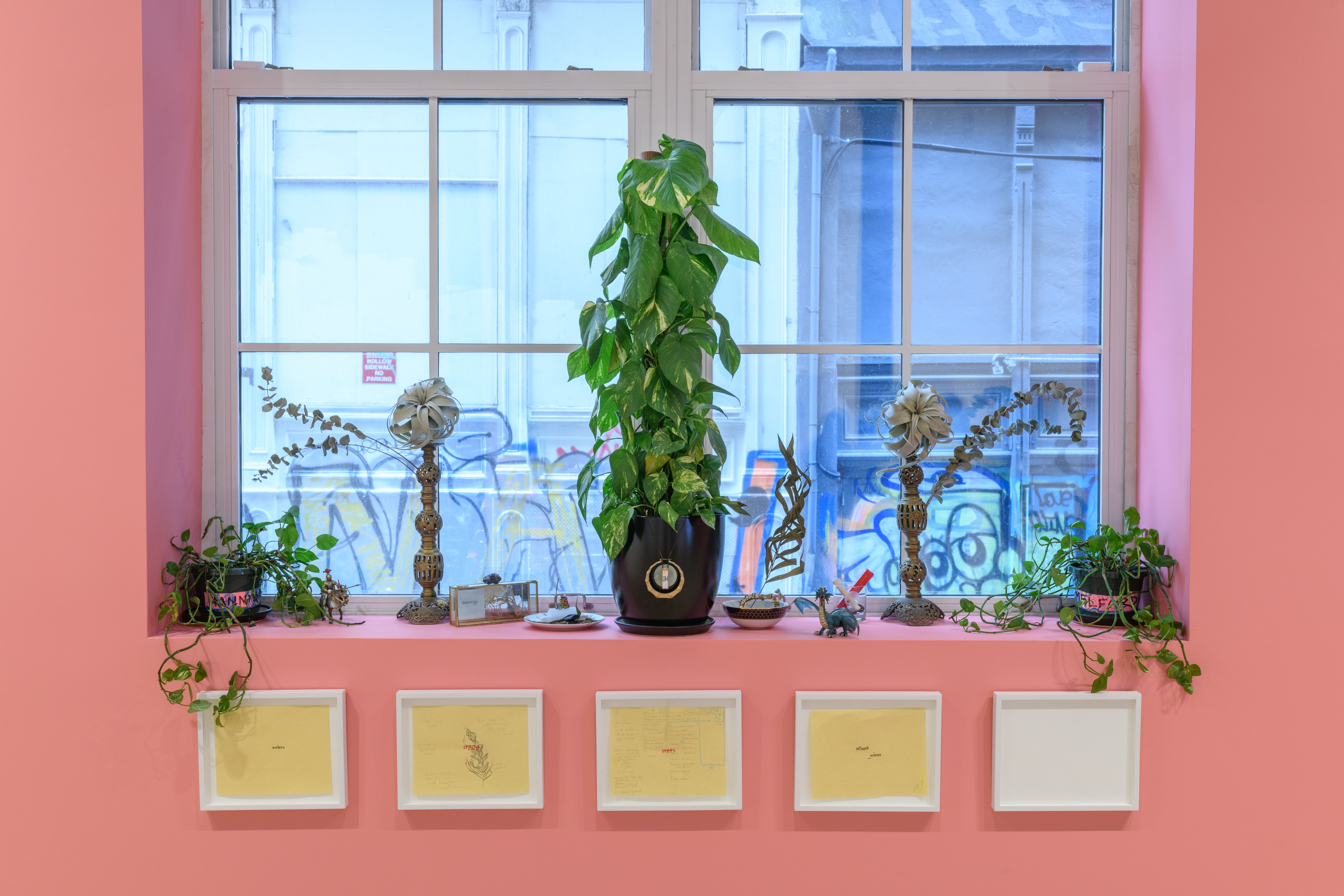 Potted plants and small figurines line the windowsill. Beneath are four framed works on paper and an empty frame.