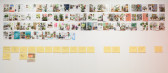 Magazine pages are arranged in several rows on a white gallery wall. Below the pages is a row of sketches on yellow paper.