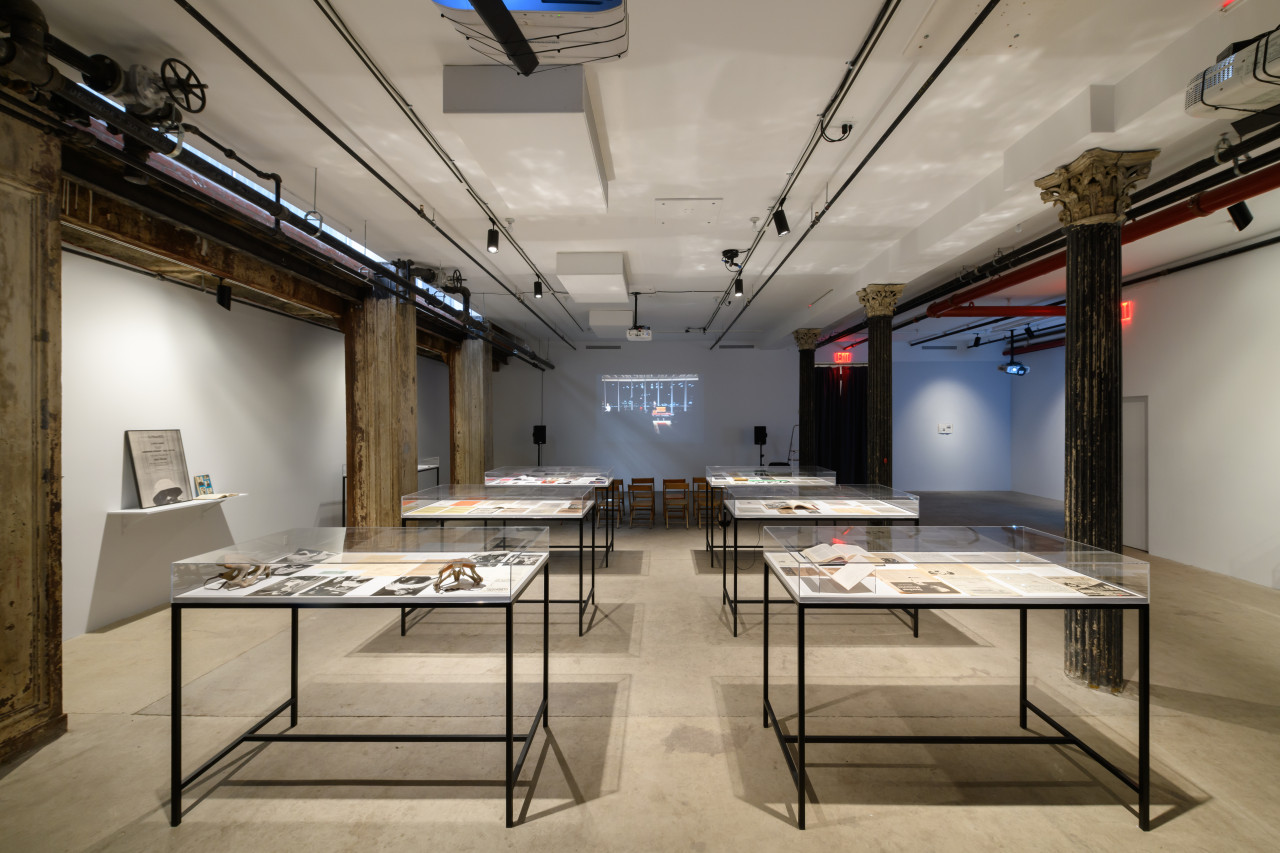 Six glass vitrines displaying books, photographs, and other paper ephemera are installed in the center of a gallery space. A film is being projected on the back wall of the exhibition space.