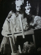 Two people in white face paint and fanciful dresses crowd around a crudely constructed wooden desk that sits off-kilter. One sits at the desk and applies makeup while the other stands to their left, gazing beyond the image.