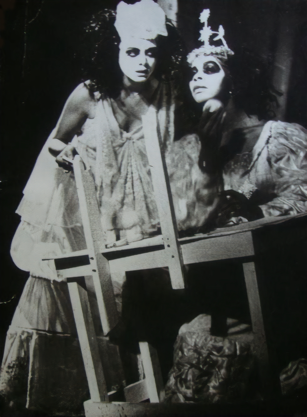 Two people in white face paint and fanciful dresses crowd around a crudely constructed wooden desk that sits off-kilter. One sits at the desk and applies makeup while the other stands to their left, gazing beyond the image.