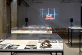 Three glass vitrines displaying books, photographs, and other paper ephemera in the center of a gallery space. A film is being projected on the back wall of the exhibition space.