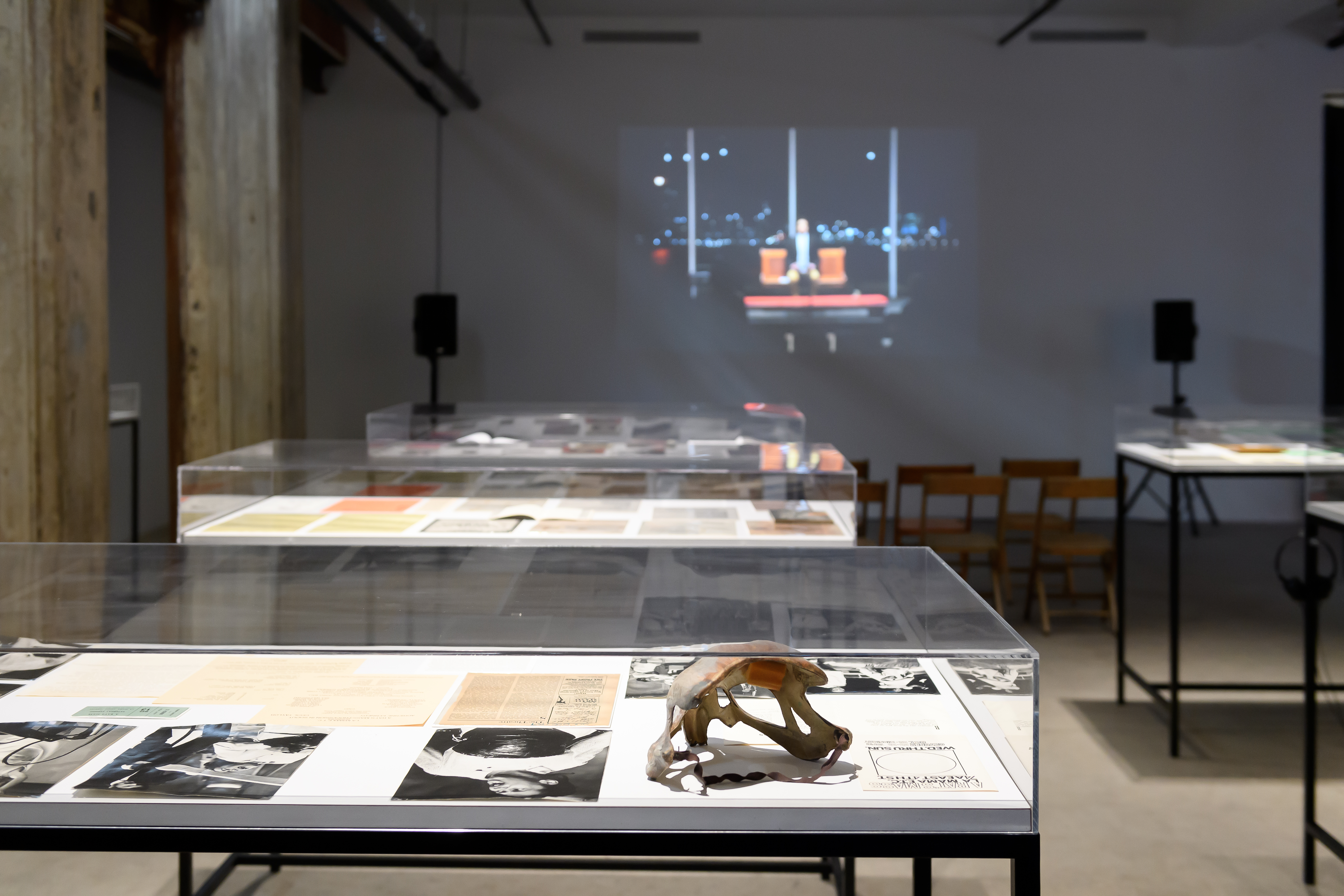 Three glass vitrines displaying books, photographs, and other paper ephemera in the center of a gallery space. A film is being projected on the back wall of the exhibition space.