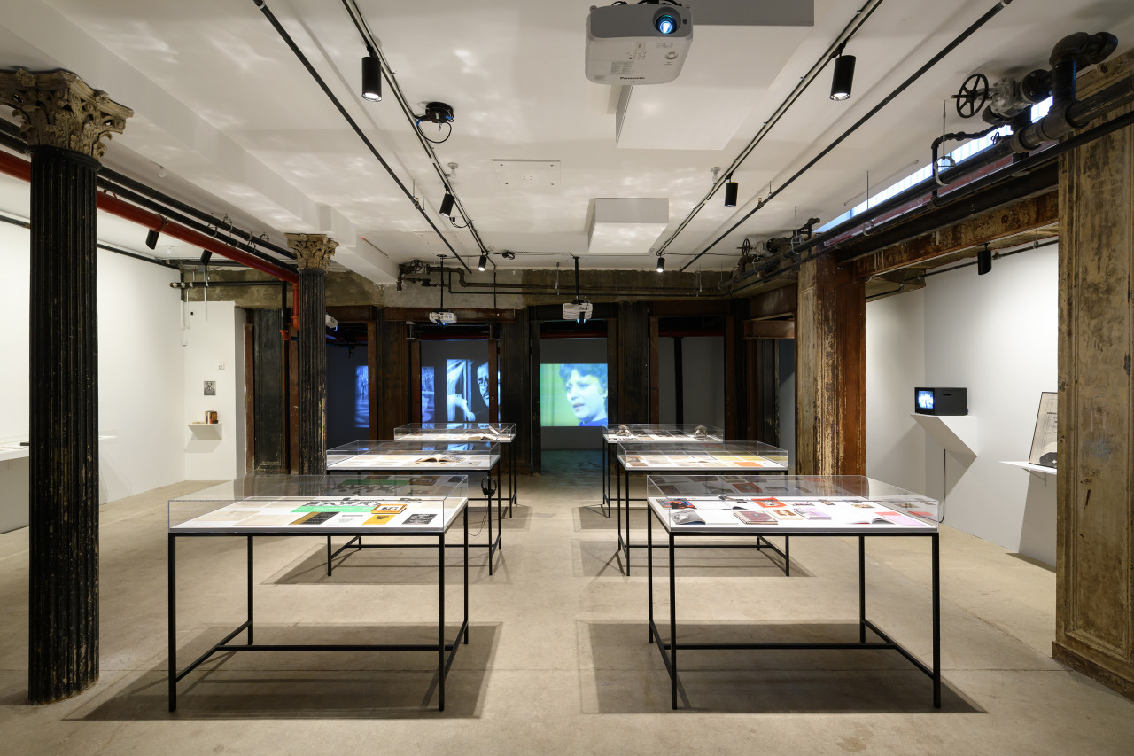 Six glass vitrines displaying books, photographs, and other paper ephemera are installed in the center of a gallery space. A series of old Hollywood films are projected on the back wall of the exhibition space.