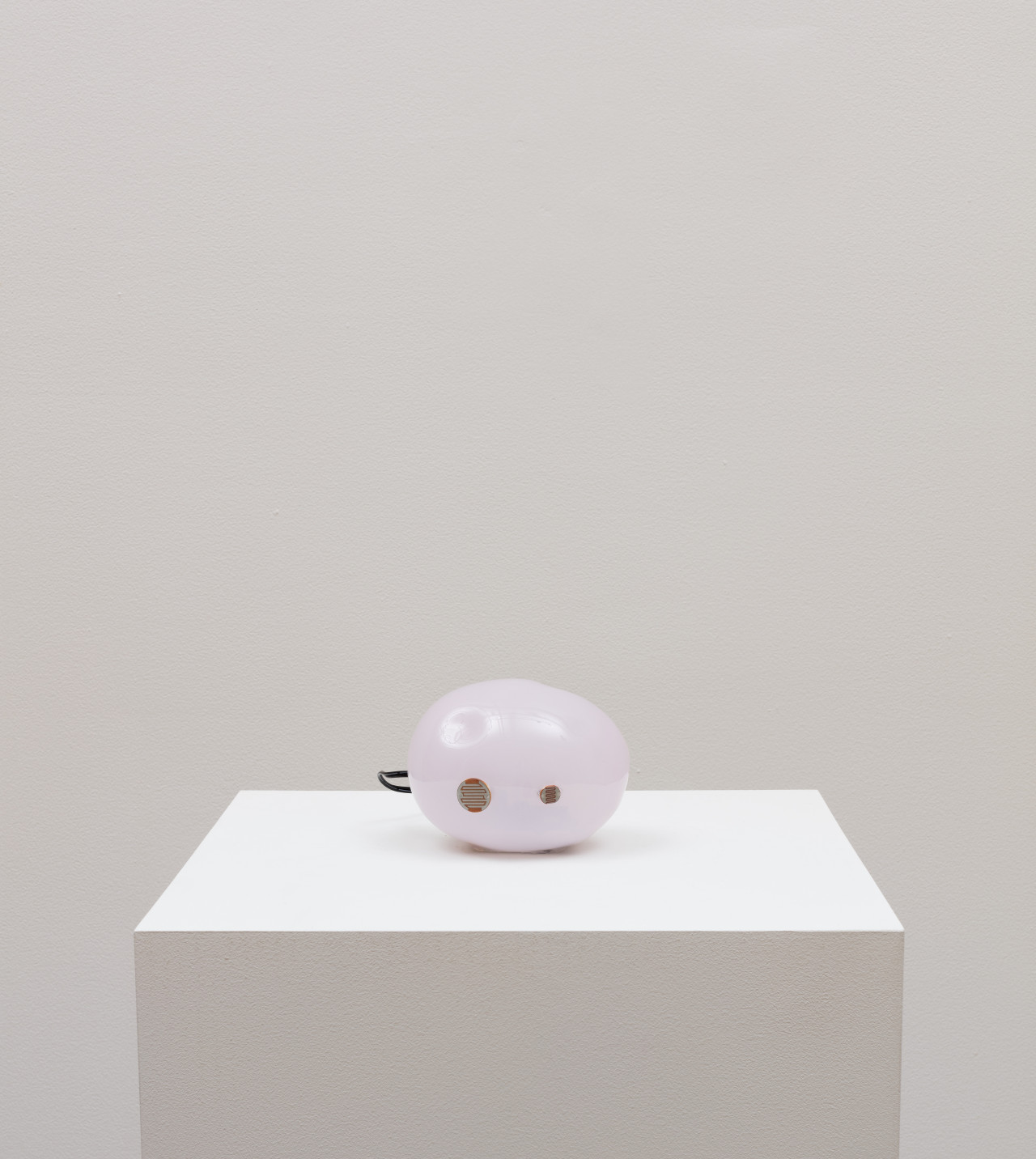 A bulbous light-hued pink sphere-like object rests upon a white plinth. When in use, a red light on the object blinks to signal that the object is functioning correctly.