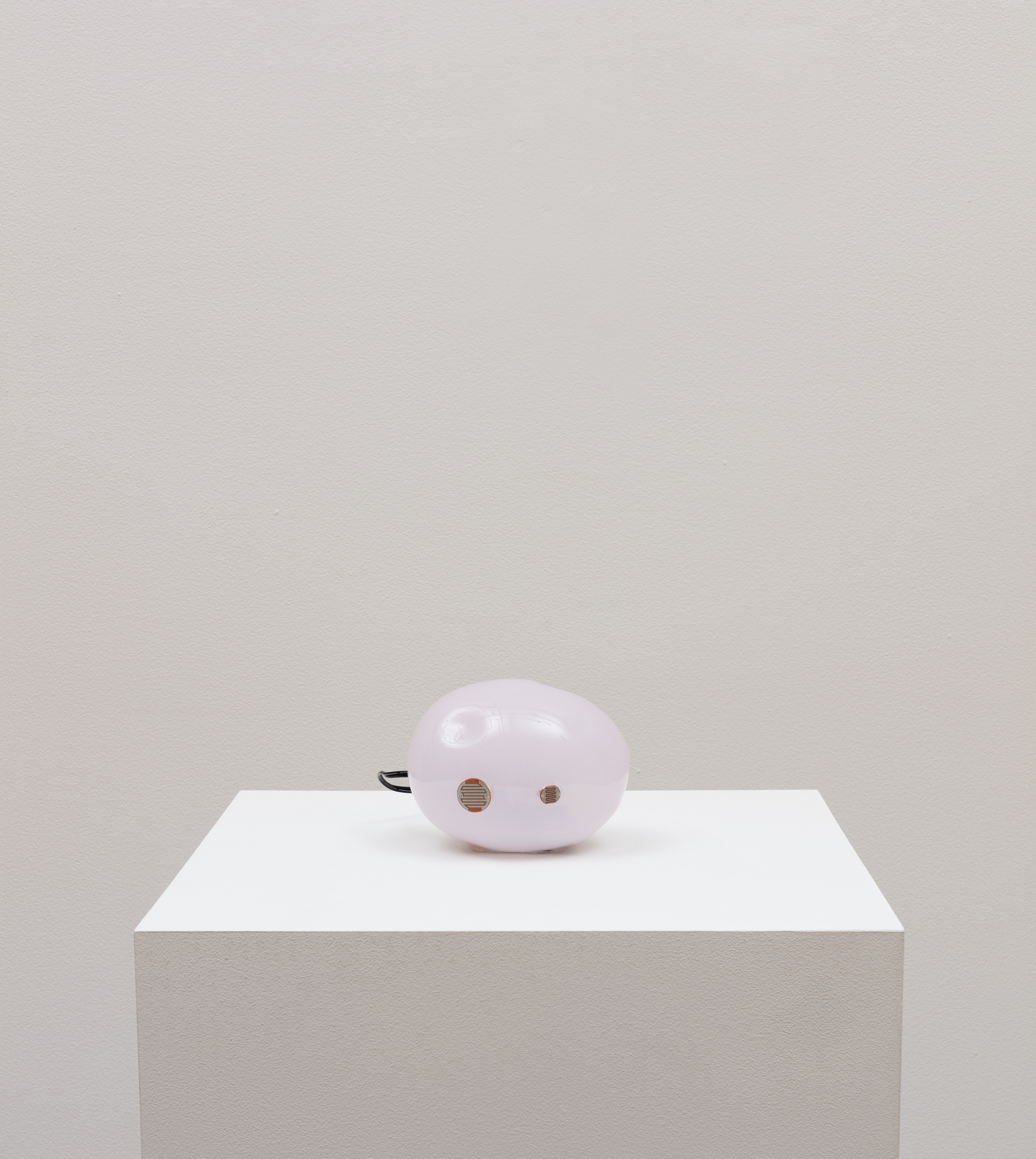 A bulbous light-hued pink sphere-like object rests upon a white plinth. When in use, a red light on the object blinks to signal that the object is functioning correctly.