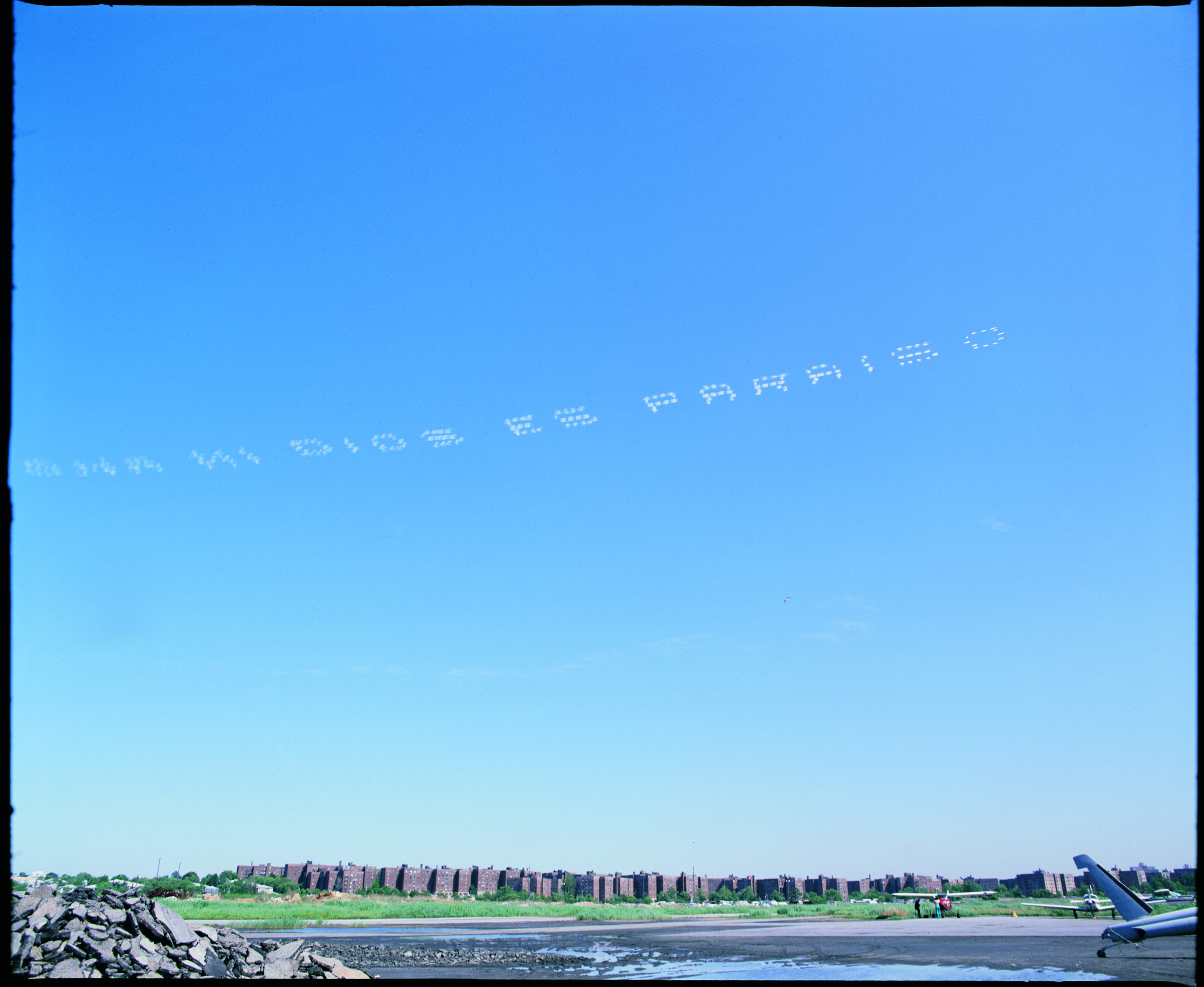 Blotted smoke-dots curve across a vast blue sky, spelling out the phrase 