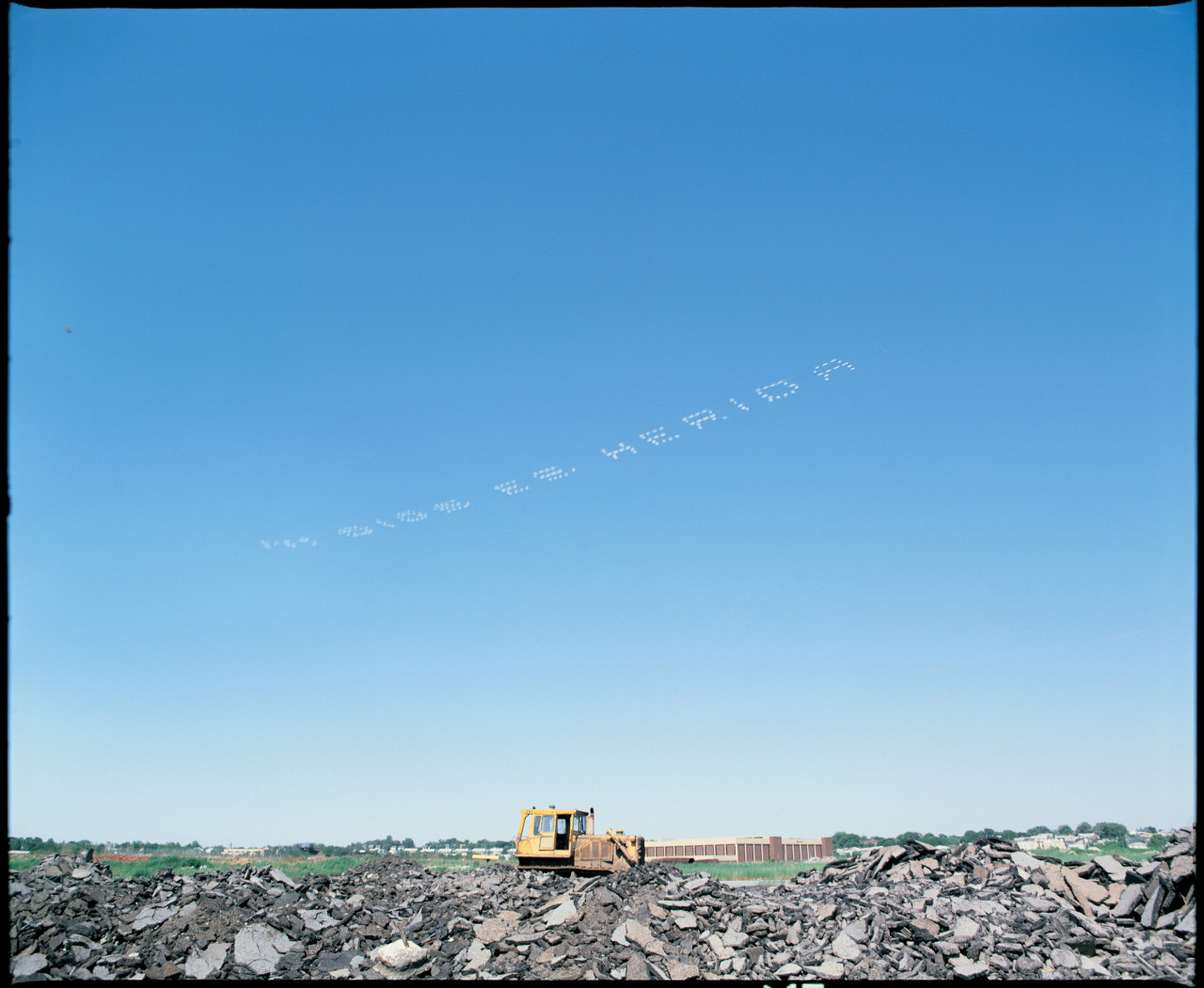 A yellow industrial machine rests atop a pile of rubble and stone. Across the clear blue sky, sky-typers write “MI DIOS ES MERIDA” in white smoke-dots.