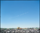 A yellow industrial machine rests atop a pile of rubble and stone. Across the clear blue sky, sky-typers write “MI DIOS ES MERIDA” in white smoke-dots.